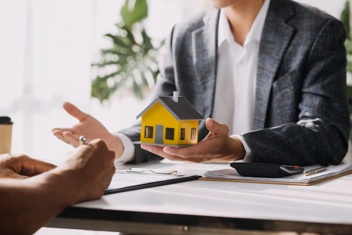 Personne en veste à carreaux présentant une maquette de maison au-dessus d’un bureau couvert de documents
