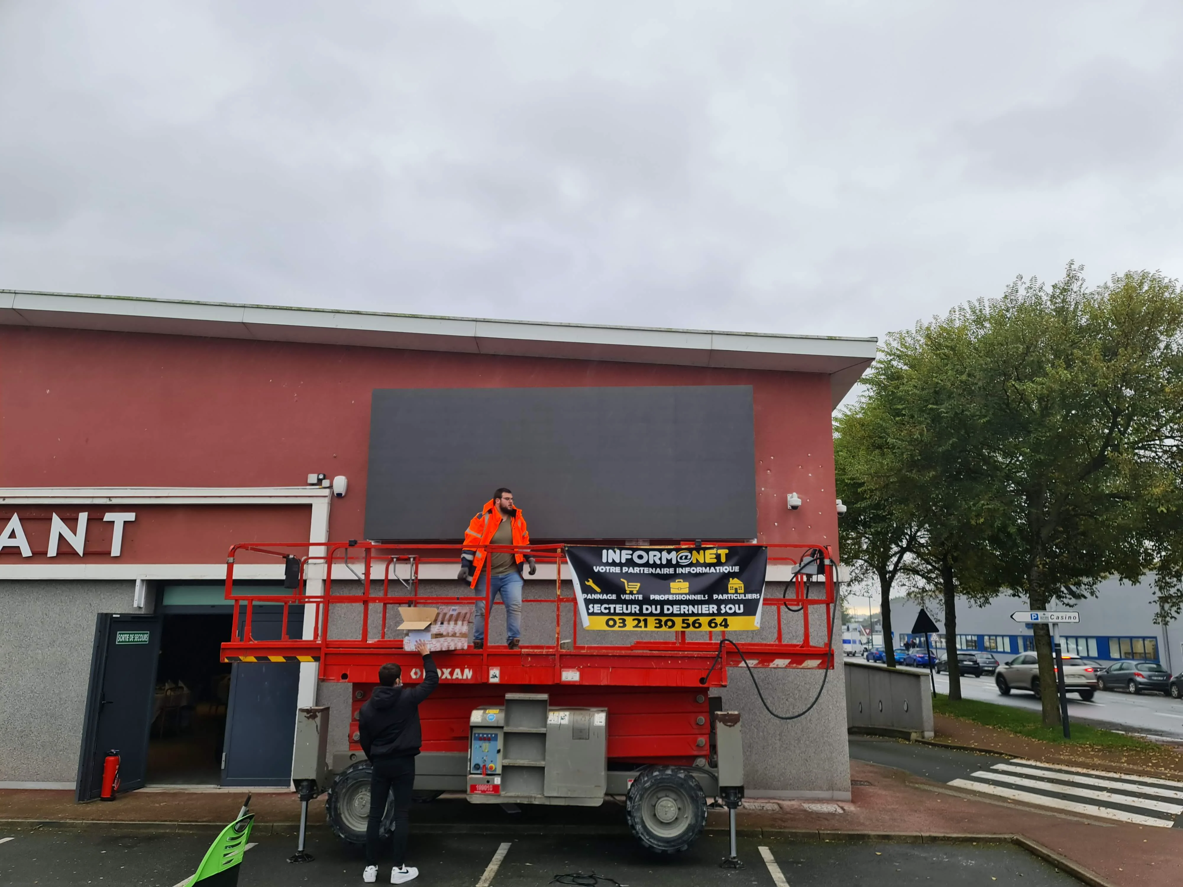 Deux personnes près d’un bâtiment commercial avec une nacelle élévatrice rouge.