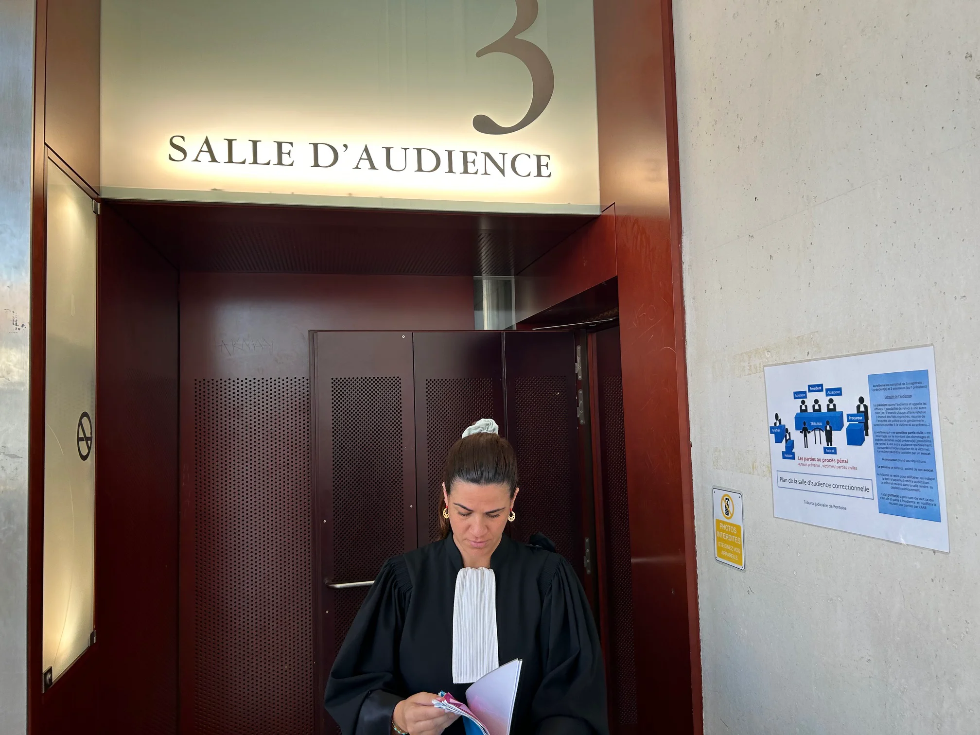 Entrée d’une salle d’audience numérotée, avec une personne en robe tenant des documents et regardant vers le bas.