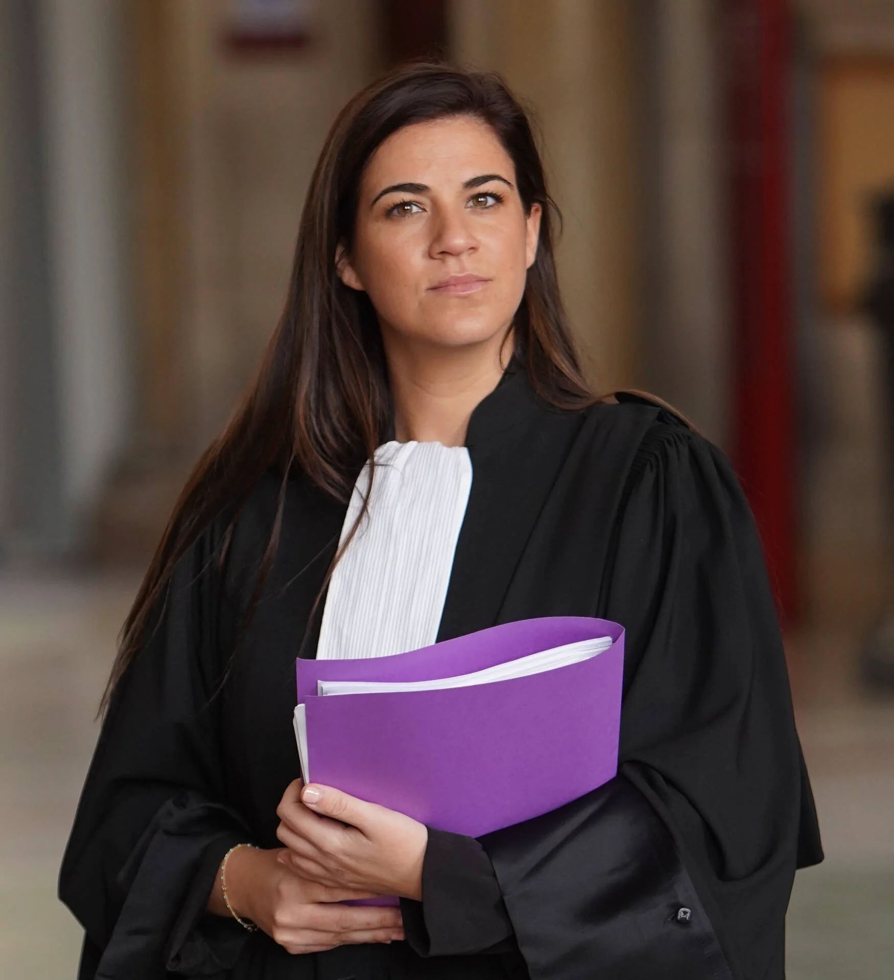 Personne en robe noire tenant un dossier violet contenant des documents blancs dans un couloir intérieur