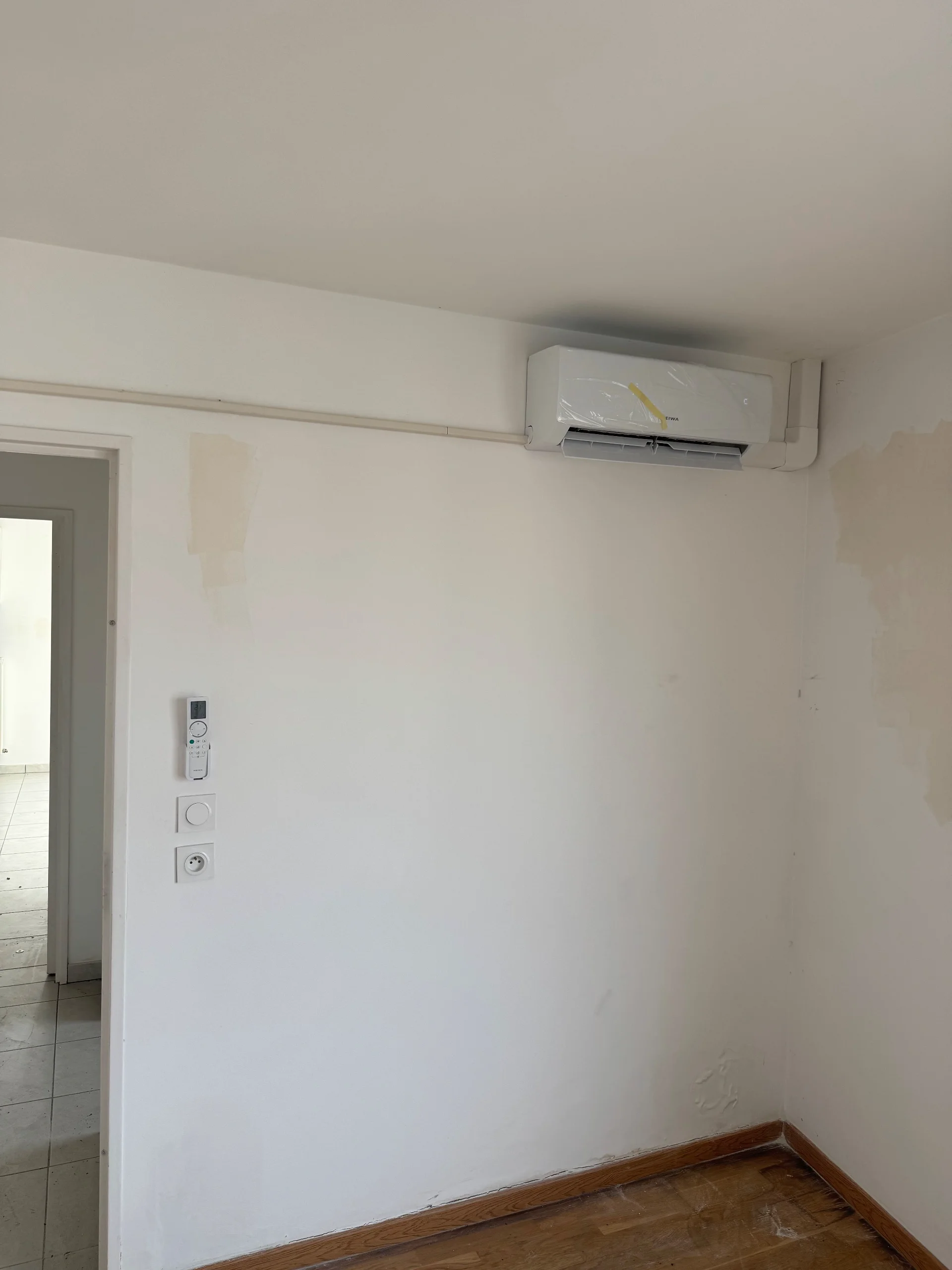 Climatiseur mural dans une pièce vide, avec une porte ouverte vers un espace carrelé.